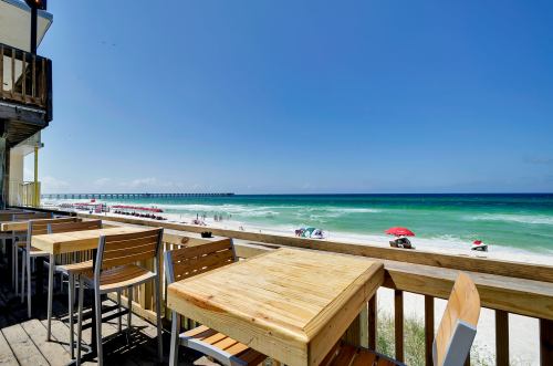 View of a beach with umbrellas, waves, and a pier, seen from a wooden deck with empty tables and chairs.