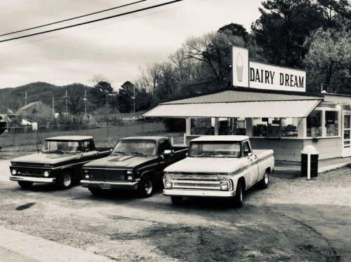 Three vintage trucks parked in front of a retro ice cream shop named "Dairy Dream" in a scenic, rural setting.