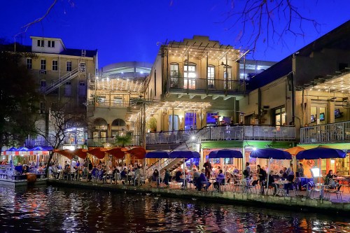A lively riverside scene at dusk, featuring outdoor dining with colorful umbrellas and illuminated buildings.