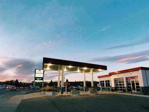 A gas station at dusk, featuring a modern canopy and a small convenience store, with colorful clouds in the sky.