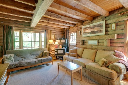 Cozy log cabin living room with wooden beams, a sofa, futon, lamps, and large windows letting in natural light.
