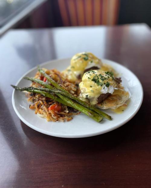 A plate with eggs Benedict on biscuits, asparagus, and a side of hash browns with vegetables, on a wooden table.