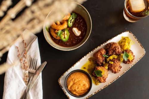 A plate of crispy fritters with sauce, a bowl of spicy shrimp, and a drink, set on a dark table with a napkin.