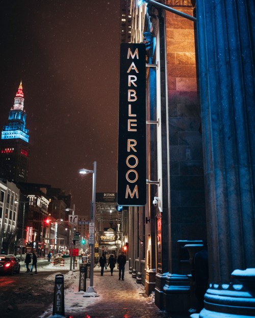 A nighttime street view of the Marble Room restaurant, illuminated by its sign, with snow falling and people walking by.