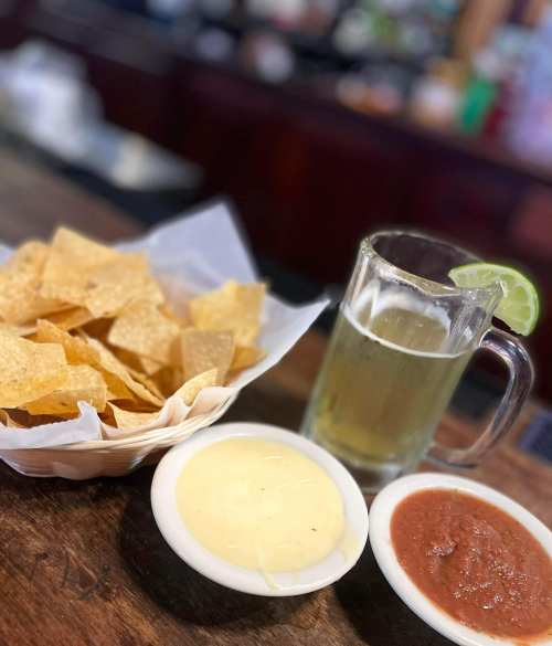 A basket of tortilla chips with cheese dip and salsa, alongside a glass of beer with a lime wedge.