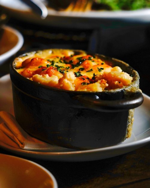 A baked dish in a black ceramic bowl, topped with golden cheese and garnished with herbs, served on a white plate.