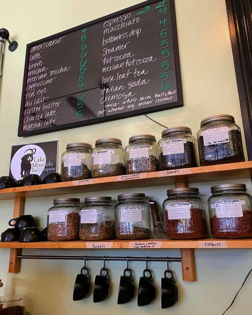 A menu board with coffee prices above jars of tea and herbs on a shelf, with black cups hanging below.