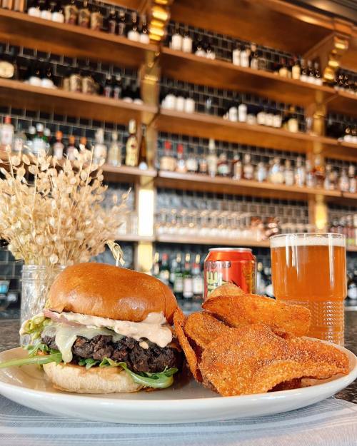 A delicious burger with lettuce and sauce, served with crispy chips, a can of soda, and a glass of beer on a bar counter.