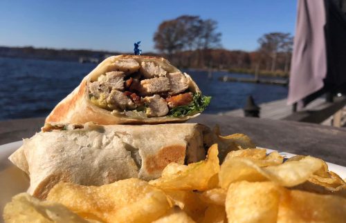 A close-up of a wrap filled with meat and vegetables, served with a side of crispy potato chips by a lake.