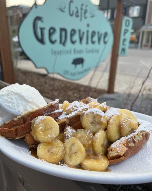 A plate of waffles topped with sliced bananas, syrup, and powdered sugar, served with a side of whipped cream.