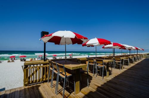 Beachside view with red and white umbrellas, wooden deck, and ocean waves under a clear blue sky.