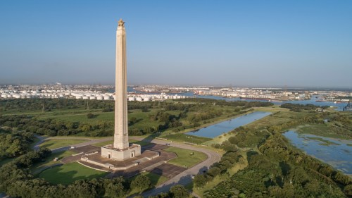 Aerial view of a tall monument surrounded by greenery and water, with a city skyline in the background.