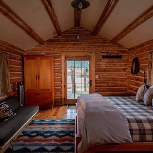 Cozy log cabin interior with wooden walls, a bed, a sofa, and a door leading to an outdoor view.