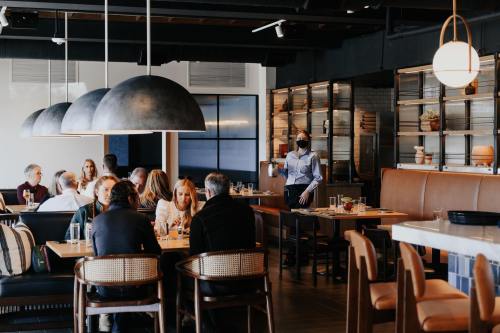 A busy restaurant interior with diners at tables and a masked server taking an order. Modern decor and warm lighting.
