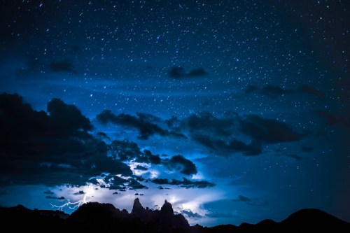 A dramatic night sky filled with stars and clouds, illuminated by distant lightning over dark mountain silhouettes.