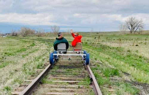 Two people ride a rail bike on an empty track, waving, with grassy fields and cloudy skies in the background.