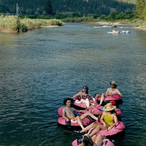 A group of people relax on pink inflatable rafts in a river, surrounded by greenery and distant hills.