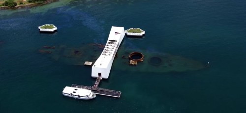 Aerial view of a white memorial structure over a submerged shipwreck, surrounded by water and small platforms.