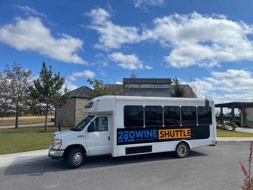 A white shuttle bus with "290 Wine Shuttle" branding parked near a building under a partly cloudy sky.