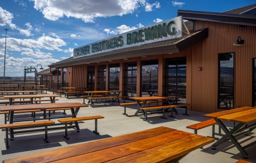 Outdoor seating area at a brewery with wooden tables and a sign that reads "Counter Brothers Brewing" under a blue sky.