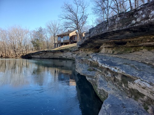 A rocky riverbank with a house perched above, surrounded by bare trees and clear blue water.