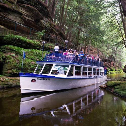 A tour boat with passengers glides through a lush, green canyon surrounded by tall trees and rocky cliffs.