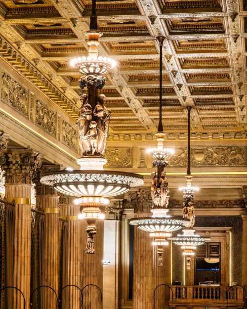 Ornate chandeliers with sculptures hang from a beautifully detailed ceiling in a grand interior space.