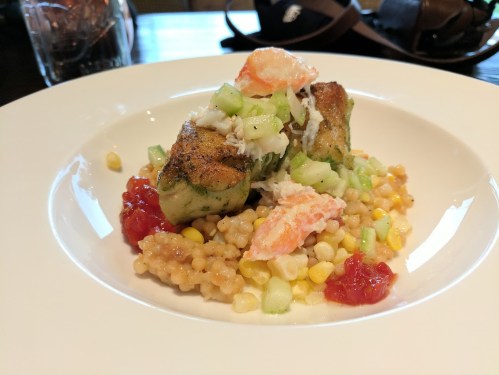 A plate of grilled fish atop a bed of couscous, corn, and diced vegetables, garnished with sauce and herbs.