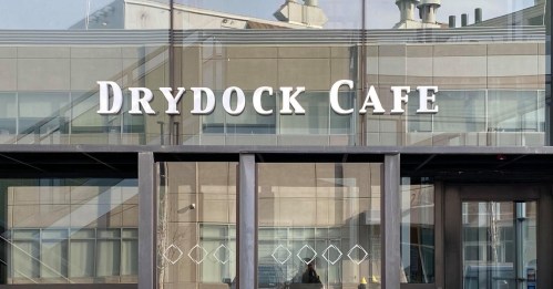Sign for "Drydock Cafe" displayed on a glass storefront with reflections of nearby buildings.