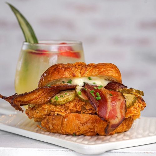 A crispy fried chicken sandwich with bacon, pickles, and mayo on a croissant, served with a refreshing drink.