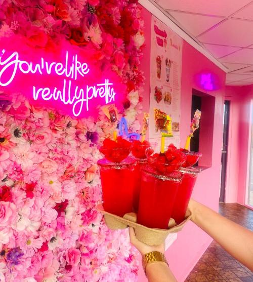 A hand holds a tray of four red drinks with raspberries, in front of a pink floral wall and neon sign.