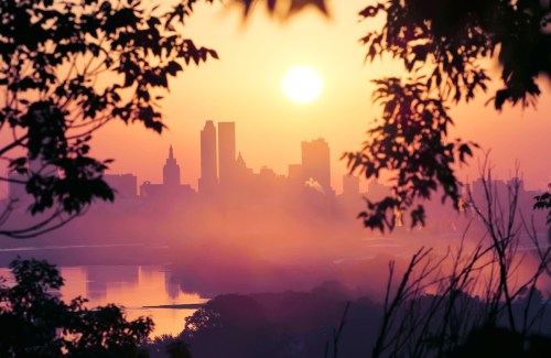 Sunset over a city skyline, framed by trees, with a hazy reflection on the water below.