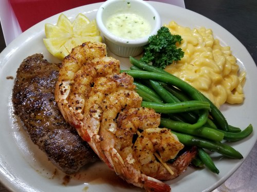 A plate featuring grilled shrimp, steak, green beans, macaroni and cheese, and lemon wedges with a dipping sauce.