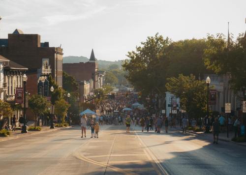 A bustling street scene with people walking, tents set up, and buildings lining the road under a warm, sunny sky.