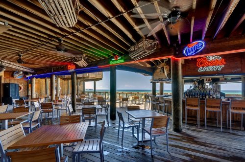 A beachside bar with wooden tables, chairs, and neon signs, overlooking the ocean through open windows.