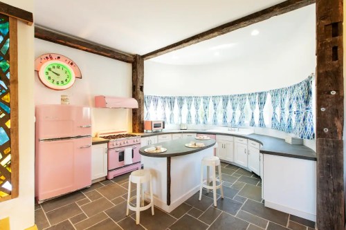 A retro-style kitchen with a pink fridge, stove, and a curved countertop, featuring blue patterned curtains and a vintage clock.