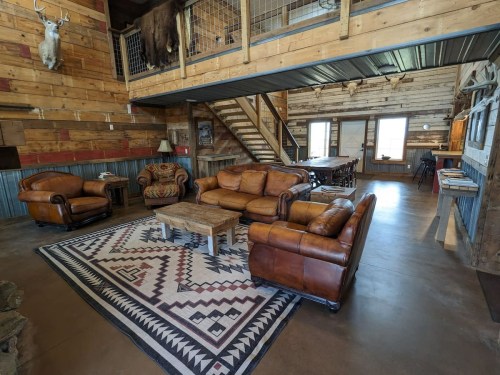 Cozy rustic living area with leather sofas, a wooden coffee table, and a staircase, featuring wood-paneled walls and a rug.