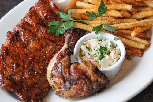 A plate featuring grilled ribs, a piece of chicken, fries, and a small bowl of coleslaw, garnished with parsley.