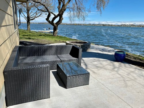 A patio with a black wicker sofa and table overlooking a lake, surrounded by trees and a clear blue sky.