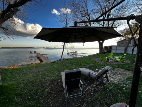 A serene lakeside view with a patio, lounge chairs, and a dock under a blue sky with scattered clouds.