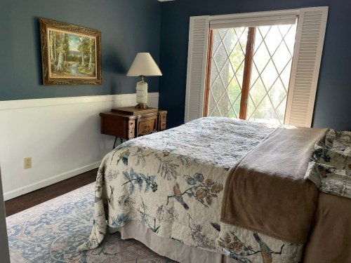 Cozy bedroom with a patterned bedspread, a lamp, and a window featuring diamond-shaped panes, framed by white shutters.
