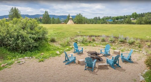 A circular seating area with blue chairs around a fire pit, surrounded by greenery and distant hills.