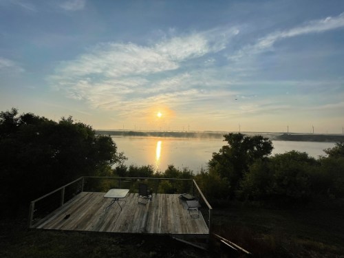 Sunrise over a calm lake, reflecting light, with a wooden deck and chairs in the foreground surrounded by greenery.