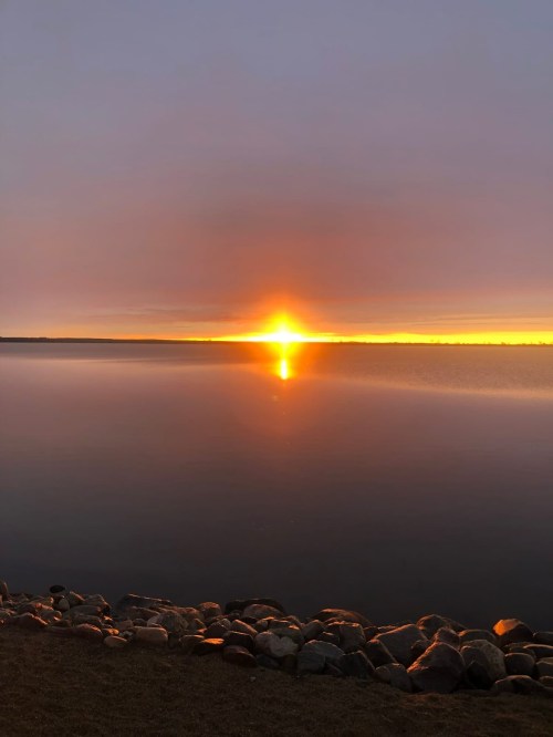 A serene lake at sunset, with vibrant orange and yellow hues reflecting on the calm water and rocky shoreline.