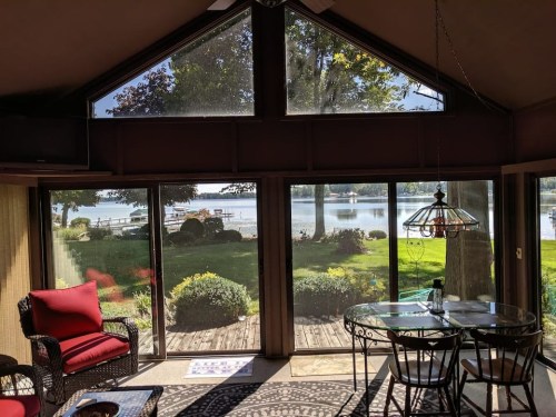 Cozy sunroom with red chairs and a glass table, overlooking a serene lake and lush greenery outside.