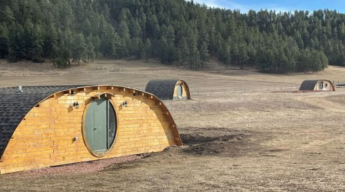 Three wooden, dome-shaped cabins are set in a grassy field surrounded by trees and a fence in the background.