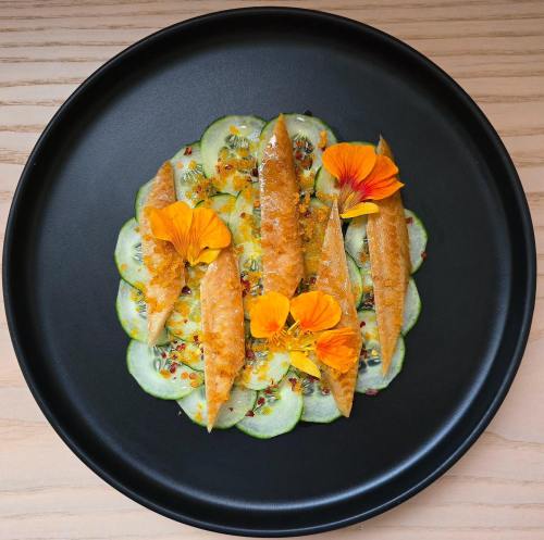 A black plate featuring cucumber slices topped with golden fish fillets and vibrant orange edible flowers.