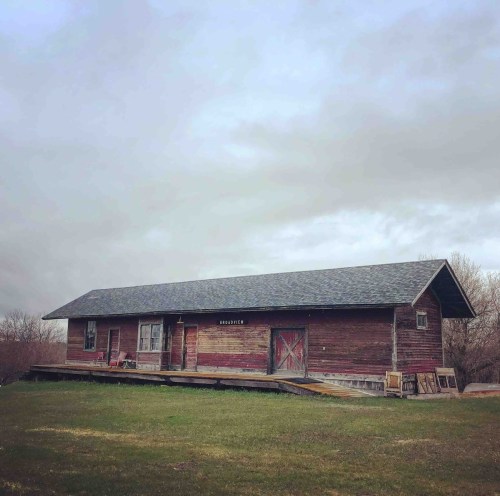 A rustic wooden building with a sloped roof, set against a cloudy sky and grassy landscape.