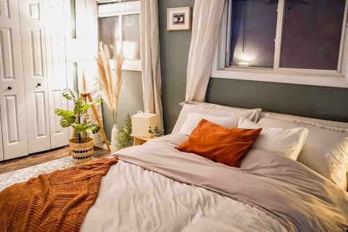 Cozy bedroom with a bed, decorative pillows, a plant, and soft lighting, featuring warm tones and a serene atmosphere.