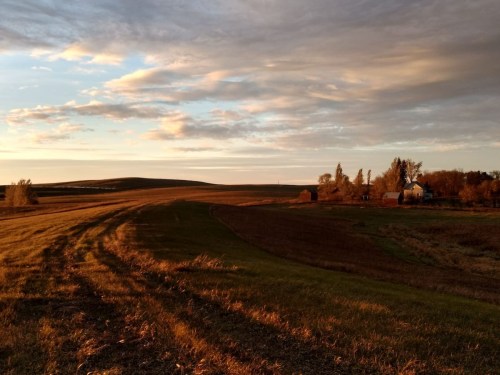 A serene landscape at sunset, featuring rolling fields, a distant farmhouse, and a colorful sky with scattered clouds.